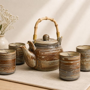 May include: A ceramic tea set consisting of a teapot with a bamboo handle and four matching cylindrical cups. The pieces feature a textured, horizontal glaze in shades of brown, grey, and cream. A small, speckled cream vase is positioned nearby.