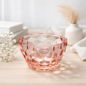 Puede incluir: Un cuenco de vidrio rosa con un patrón geométrico de diamantes. El cuenco está sobre una superficie de madera clara. El cuenco está vacío y parece ser un objeto decorativo. El fondo incluye flores blancas, un jarrón blanco y un paño blanco.