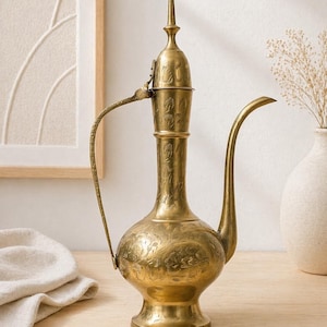Può includere: Brocca vintage in ottone con beccuccio e manico allungati. La brocca presenta intricate incisioni floreali e una parte superiore a punta. È esposta su una superficie di legno, mettendo in mostra il suo design ornato e l'estetica antica.