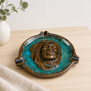 May include: A round ceramic ashtray featuring a raised, brown, sculpted relief of a person wearing a headdress in the center. The basin is glazed in a vibrant turquoise blue, while the outer rim and the central figure are finished in a glossy brown.