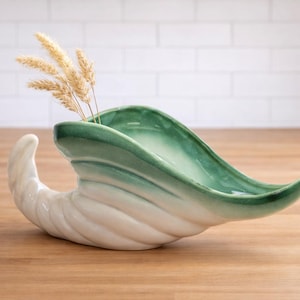 May include: A ceramic decorative vase in a seashell shape, with a white and green gradient finish. The vase holds a few stalks of dried wheat. The vase is placed on a wooden surface.