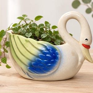 Peut inclure: Un cache-pot en céramique en forme de cygne au corps crème, doté d'un motif d'aile peint dans des tons dégradés de vert clair et de bleu vif. Le cygne possède un bec rouge et des yeux noirs, et contient une petite plante verte feuillue.