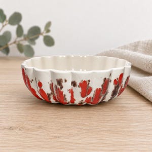 Peut inclure: Un bol en céramique peu profond et rond avec un bord festonné. L'extérieur présente une base de couleur crème avec un motif décoratif composé d'éclaboussures de peinture rouge vif et brun foncé. L'intérieur du bol est d'un crème uni et lisse.