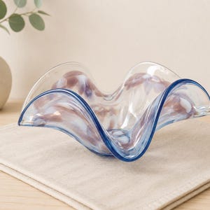 Puede incluir: Un cuenco de cristal transparente con un borde ondulado y fruncido que presenta patrones de remolinos en tonos azul claro y violeta suave. El cuenco tiene una calidad translúcida, permitiendo que la luz pase a través de su forma orgánica.