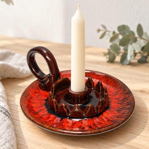 May include: A round ceramic candle holder with a glossy red and black glaze, featuring a central socket for a single taper candle and a curved handle on the side. The base has a raised, scalloped rim surrounding the central candle holder section.