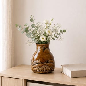 May include: A brown ceramic vase with a textured, carved design, holding a bouquet of white flowers and green foliage. The vase sits on a light wood surface, next to a closed book. The overall aesthetic is natural and minimalist.