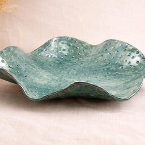 Puede incluir: Un cuenco de cerámica turquesa con un borde ondulado y una superficie texturizada con pequeñas protuberancias en relieve. El cuenco se exhibe sobre una superficie blanca, con un jarrón blanco y plantas secas en el fondo.