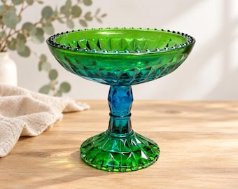 Puede incluir: Un cuenco con pedestal hecho de vidrio verde vibrante con un patrón de diamantes facetados en el exterior y un borde con cuentas. El tallo cambia a un tono azul profundo, sosteniendo el cuenco ancho y poco profundo sobre una base circular.