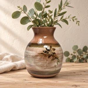 May include: A round ceramic vase with a glossy finish, featuring horizontal bands of brown, tan, and muted green tones. The body of the vase is decorated with a hand-painted dark brown floral motif. The vase contains several green eucalyptus stems.