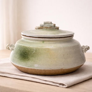Puede incluir: Una olla de cocina de cerámica hecha a mano con tapa. La olla es de color verde claro con una base marrón y una superficie texturizada. Tiene dos asas decorativas y una tapa escalonada. La olla está sobre una servilleta de lino beige.