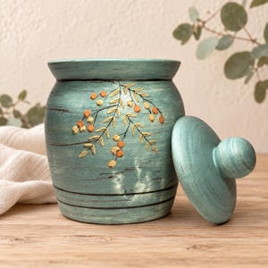 May include: A teal wooden canister with a removable lid, featuring a hand-painted design of delicate branches with small orange berries and thin black horizontal lines encircling the body of the container, showcasing a weathered finish.