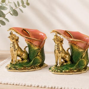 May include: A pair of decorative figurines featuring a metallic gold dog sitting beside a large, pink and green calla lily flower. The dog is looking upward with its mouth open, and the flower serves as a hollow vessel with a glossy finish.