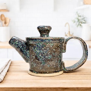 Puede incluir: Una tetera de cerámica moteada con una base marrón oscuro y un diseño moteado azul y verde. La tetera tiene un asa curva y un pico, y se asienta sobre una superficie de madera. La tapa tiene un pequeño pomo.