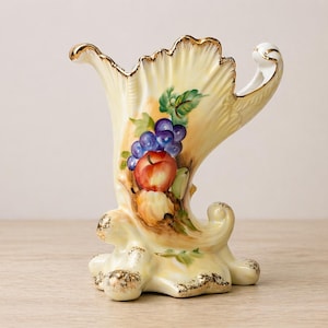 Puede incluir: Un jarrón de cerámica color crema con ribete dorado y diseño de frutas. El jarrón presenta un racimo de uvas moradas, una manzana roja y otras frutas. El jarrón tiene una forma curva única y se asienta sobre una base de tres patas.