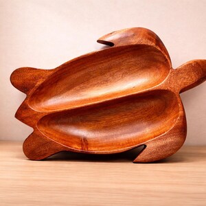 May include: A handcrafted wooden turtle-shaped serving dish. The dish is made of a rich, reddish-brown wood and features two separate compartments. The turtle's head and flippers are incorporated into the design, creating a unique and decorative piece.