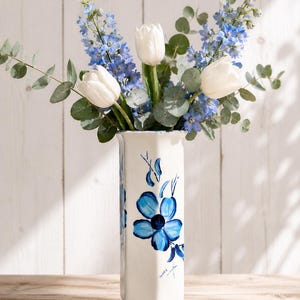 Puede incluir: Un jarrón de cerámica blanca con un diseño floral azul contiene un ramo de tulipanes blancos, delfinios azules y hojas de eucalipto verdes. El jarrón tiene forma rectangular con una base acampanada y un borde ligeramente festoneado.