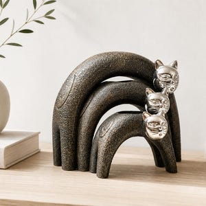 Op de afbeelding: Een decoratieve sculptuur met drie gestileerde kattenfiguren die op elkaar zijn gestapeld. De katten hebben een getextureerd, donker bronzen lichaam en zilverkleurige hoofden. De sculptuur staat op een lichtgekleurd houten oppervlak.