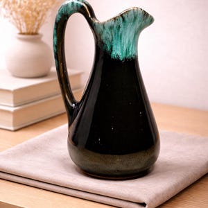 Puede incluir: Jarra de cerámica negra con asa curva y pico vertedor. La parte superior y el asa son de color verde turquesa. La jarra está sobre un paño beige en una mesa de madera. Un jarrón con flores secas está al fondo.