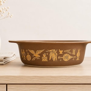 May include: A brown casserole dish with gold-colored decorative elements. The dish features a series of gold illustrations, including an eagle, corn, and other vintage-style designs. The dish is on a wooden surface.