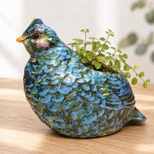 May include: A bird-shaped ceramic planter with a glossy finish in shades of blue, teal, and yellow. The textured surface mimics feathers, and the piece features a small orange beak. A small green plant with rounded leaves grows from the opening.
