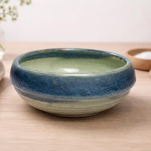 Puede incluir: Un cuenco de cerámica hecho a mano con un esmalte azul y verde. El cuenco tiene una forma redondeada y una superficie texturizada. El interior es verde claro, mientras que el borde exterior es azul oscuro. El cuenco está sobre una superficie de madera.