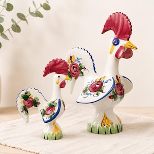 Puede incluir: Dos figuras de gallo de cerámica blanca con motivos florales pintados a mano en rojo, verde y azul. Cada gallo tiene una cresta roja prominente, un pico amarillo y una base decorada con trazos verdes, sobre una superficie plana.