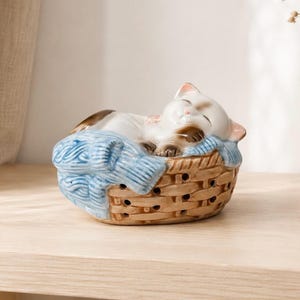 May include: A ceramic figurine of a sleeping cat with brown and white markings, resting inside a brown woven basket. The cat is curled up on a light blue textured blanket that drapes over the edges of the basket, which features small cutouts.