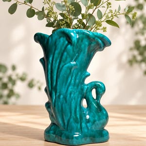 May include: A glossy teal ceramic vase shaped like a swan with a flared, textured opening. The body of the vase features a deep, mottled turquoise glaze with darker highlights. Green eucalyptus branches are placed inside the swan-shaped vessel.
