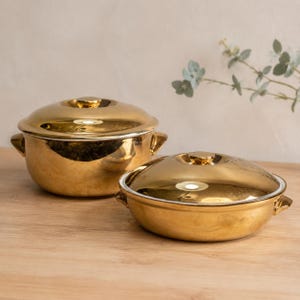 Könnte beinhalten: Zwei metallisch goldene Keramik-Servierschalen mit passenden Deckeln und seitlichen Griffen. Eine Schale ist tief und abgerundet, die andere flach und breit. Beide Stücke haben eine hochglänzende, polierte Goldoberfläche und glatte, geschwungene Kanten.