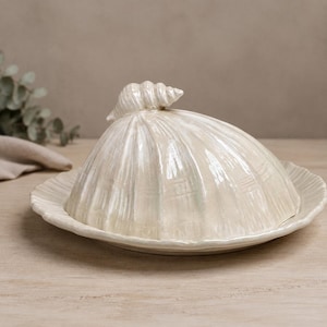 Puede incluir: Una mantequillera de cerámica color crema con una tapa abovedada que presenta un asa esculpida en forma de concha marina. La tapa y la base circular a juego tienen una textura acanalada con un acabado brillante y un diseño cohesivo.