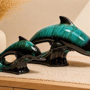 May include: Two ceramic dolphin sculptures. The dolphins are teal and black, with a glossy finish. The larger dolphin is arched, and the smaller dolphin is leaping. The sculptures are displayed on a wooden surface.