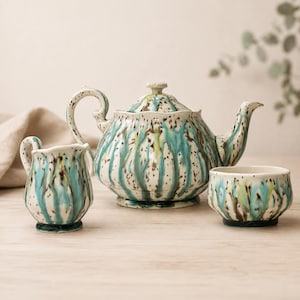 May include: A ceramic teapot with a lid, a small creamer jug, and a tea cup. Each piece features a cream-coloured base with vertical turquoise blue drips and dark brown speckles throughout the glazed surface of the pottery set.