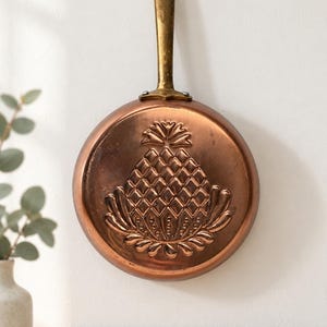 Puede incluir: Una sartén redonda de cobre con un diseño de piña en relieve en su base. La sartén tiene un mango largo de tono latón desgastado fijado en la parte superior, diseñado para colgar. La superficie metálica muestra un acabado marrón rojizo.