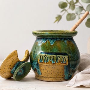 Op de afbeelding: Een ronde keramische honingpot met een gespikkelde gele basis en een glanzend groen en blauw druipglazuur. Het woord HONEY staat in reliëf op de voorzijde. Een bijpassend gespikkeld geel deksel ligt links en een lichte houten honingdrijver zit erin.