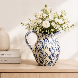 Puede incluir: Una jarra crema con un patrón de salpicaduras azul, llena de flores blancas. La jarra tiene un asa curva y está sobre una superficie de madera. Al lado, dos libros con los títulos "THE KINFOLK HOME" y "THIS IS HOME".