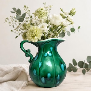 May include: A glossy emerald green ceramic pitcher with a rounded body, vertical ribbed detailing, and a decorative scrolled handle. The pitcher is filled with a bouquet of white and pale green flowers, including roses and eucalyptus sprigs.