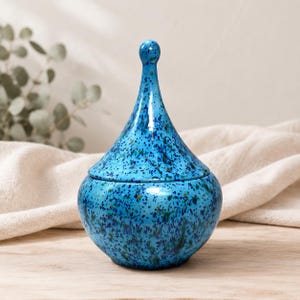 May include: A ceramic jar with a rounded base and a tall, pointed lid. The surface features a vibrant blue glaze with a speckled pattern of dark blue and green dots throughout. The object has a smooth, glossy finish and a distinct teardrop shape.