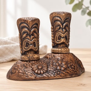 May include: A brown salt and pepper shaker set featuring two vertical tiki-style figures with carved facial expressions. The shakers are mounted on a shared oval base decorated with a relief pattern of various flowers and foliage in a dark finish.