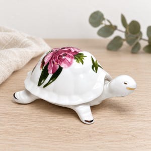 Puede incluir: Una figura de tortuga de cerámica blanca con un patrón floral detallado en su caparazón, que presenta flores de color rosa vibrante y hojas verdes. La tortuga tiene un acabado liso y brillante, un pequeño pico pintado de oro y extremidades.