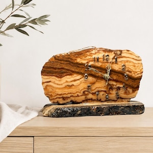 May include: A horizontal table clock crafted from a slice of natural wood with prominent brown and tan grain patterns. The clock face features metallic numerals and thin hands, mounted on a dark, rough-edged wooden base with a natural bark texture.