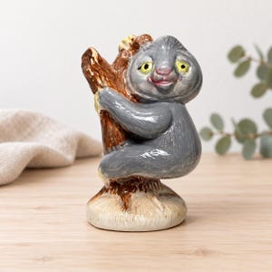 May include: A glossy ceramic figurine of a grey sloth clinging to a brown textured tree trunk. The sloth has yellow eyes, a pinkish nose, and a small smile. The base is a light beige colour, and the entire piece has a smooth, reflective finish.