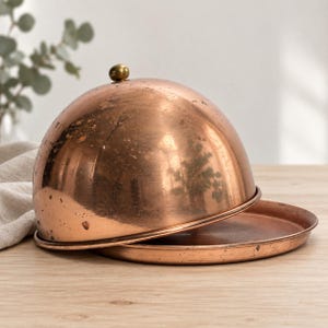 May include: A polished copper food dome with a rounded shape and a small brass knob handle on top, resting on a matching circular copper tray. The metal surface shows a warm, metallic finish with subtle signs of wear and natural patina.