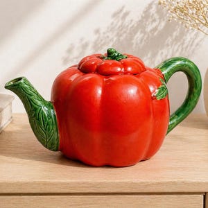 May include: A ceramic teapot shaped like a red bell pepper with a green handle, spout, and stem. The teapot is sitting on a light-colored wooden surface. A small vase with dried flowers is in the background.