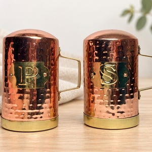 May include: A pair of cylindrical salt and pepper shakers made of hammered copper with a polished brass base and handle. Each shaker features a brass plaque on the front, with the left shaker embossed with the letter P and the right with the letter S.