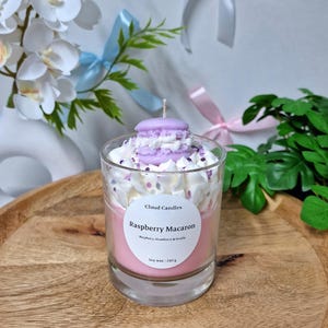 Puede incluir: Una vela de macaron de frambuesa en un vaso transparente. La vela tiene una base rosa y está cubierta con glaseado blanco y un macaron morado. La etiqueta dice "Raspberry Macaron" y "Soy wax | 240 g".