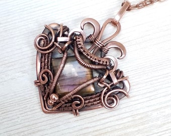May include: A handcrafted copper pendant necklace featuring a square wire-wrapped frame with a central, iridescent gemstone. The pendant has intricate wire detailing, including swirls and a fleur-de-lis design, suspended from a copper chain.