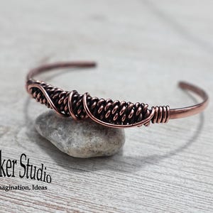 Puede incluir: Un brazalete de cobre con un diseño tejido en el centro. El brazalete descansa sobre una pequeña piedra gris. El logotipo de GemSeeker Studio es visible en la esquina inferior izquierda. Un accesorio elegante.