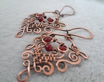 May include: A pair of copper-colored earrings with intricate wirework and small red beads. The earrings feature a swirling, open design with woven sections and are displayed on a light gray surface.
