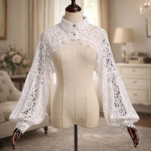 May include: A white lace bolero with a high collar and long, voluminous sleeves. The delicate floral lace design is visible throughout the garment, which is displayed on a mannequin. The bolero has button closures at the collar.