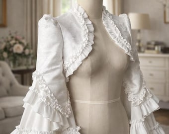 May include: A white bolero jacket with ruffled trim and bell sleeves. The jacket is displayed on a mannequin. The fabric has a subtle floral pattern. The bolero is a cropped style, perfect for layering.